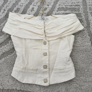 Vit korsettopp från NA-KD med knappar - Supersnygg vit korsettopp från NA-KD med bred offshoulder-krage och markerad midja. Toppen har unika fyrkantiga knappar framtill och två dekorativa fickor. Materialet är strukturerat och känns lyxigt, perfekt för att sticka ut.
