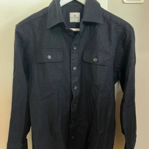 Marinblå overshirt från John Henric - Marinblå overshirt från John Henric med klassisk krage, två bröstfickor med knappar och knappstängning hela vägen. Materialet är 100% ull och har en lite borstad känsla, perfekt för lager-på-lager. Snygg och enkel att styla till jeans eller chinos. Storleken är M men skulle säga att den passar S då jag har vuxit ut den och har vanligtvis M. Hör av er om ni vill ha fler bilder