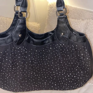 Vintage handväska  - Snygg svart axelväska med glittriga prickar och detaljer i blankt syntetmaterial. Väskan har breda axelband med silvriga spännen och ett rymligt huvudfack. Perfekt för dig som vill ha en väska som sticker ut och rymmer allt du behöver. Köpte den vintage, har använt fåtal gånger💕