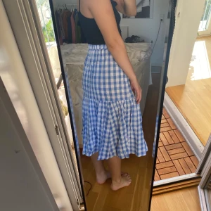 Blårutig volangkjol från Monki XS - Supersöt midilång kjol från Monki i blåvitt rutigt mönster. Kjolen har volang nertill och hög midja, perfekt för en chill och somrig look. Tillverkad i mjuk bomull som känns lätt och luftig.