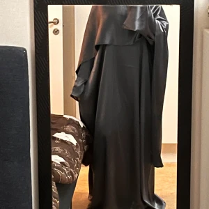 Grå/mörkblå färgad långklänning i satinlook - En grå/mörkblå långklänning med glansig satinlook och vida ärmar. Klänningen har en lös passform och ett extra lager tyg över axlarna som kan användas som sjal eller på axlarna precis som i bilden och ger en  elegant vibe. Perfekt för dig som gillar minimalistisk och stilren design.