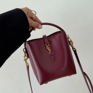 Vinröd axelväska från Saint Laurent - Snygg vinröd axelväska från Saint Laurent i slätt skinn med guldiga metalldetaljer och ikoniskt YSL-emblem framtill. Väskan har både handtag och justerbar axelrem, perfekt för en trendig look. Stilren bucket-modell som rymmer det viktigaste.