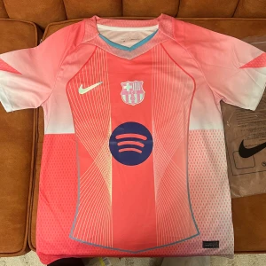 Barcelona rosa tshirt  - Säljer en FC Barcelona fotbollströja från Nike i rosa och ljusrosa med vita och blå detaljer. Tröjan har korta ärmar, klubbmärke på bröstet och Spotify-logga framtill. Tillverkad i lätt och ventilerande funktionsmaterial, otestad 