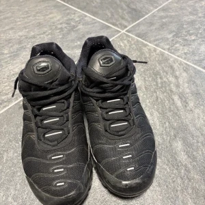 Svarta Nike Air TN - Säljer ett par helsvarta Nike Air Max sneakers med klassisk vågig design och synlig Air-sula. Skorna har snörning, mesh- och syntetmaterial samt diskreta reflekterande detaljer. Perfekta för dig som gillar streetwear och sportig stil.