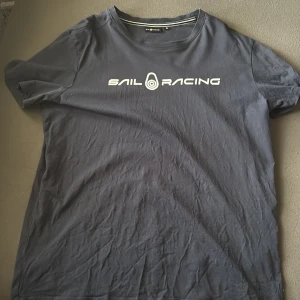 Mörkblå Sail Racing t-shirt XL - Snygg mörkblå t-shirt från Sail Racing i storlek XL. Klassisk passform med rund hals och vit logga tryckt på bröstet. Tillverkad i mjuk bomull som känns skön mot huden. Perfekt för dig som gillar stilren och sportig look.