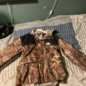 Camouflage vindjacka från The North Face - Säljer en vindjacka från The North Face i storlek M. Jackan har camouflage-mönster i bruna och svarta toner, svarta axelpartier och huva med snörning.