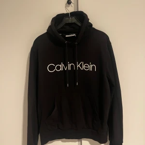 Svart Calvin Klein hoodie - Svart hoodie från Calvin Klein med stor vit logga framtill. Klassisk känguruficka, dragsko i huvan och ribbade muddar. Mjuk och skön i bomullsmix, perfekt för chill dagar. Snygg och enkel stil som funkar året runt.