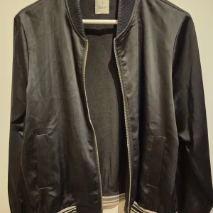 Svart bomberjacka från Culture - Svart bomberjacka från Culture med klassisk ribbad krage och muddar. Jackan har dragkedja framtill, två fickor och diskreta ränder i vitt och svart på mudden. Snygg och stilren modell i blankt material som passar perfekt till streetwear.