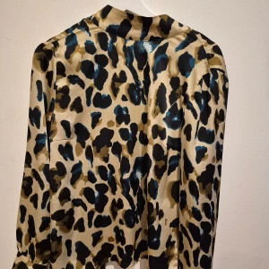 Leopardmönstrad blus från Vila - Snygg blus från Vila i leopardmönster med beige, svart och blå toner. Blusen har lång ärm och knytdetalj bak vid halsen som ger en cool touch. Materialet känns glansigt och mjukt, perfekt för dig som vill sticka ut lite extra.