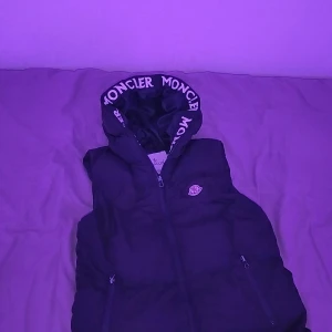 Svart dunväst från Moncler med huva - Snygg svart dunväst från Moncler med huva och dragkedja framtill. Huva med Moncler-logga tryckt runt kanten och två fickor med dragkedja. Perfekt för lager-på-lager och streetstyle.