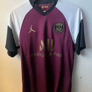 Psg t shirt - Snygg PSG fotbollströja i samarbete med Jordan. Vinröd bas med svarta och vita detaljer på ärmarna, guldiga loggor och diskreta ränder. Ikonisk Paris Saint-Germain badge på bröstet. Tillverkad i lätt och ventilerande material.