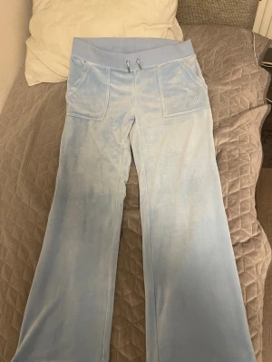 Ljusblå mjukisbyxor från Juicy Couture - Säljer ett par ljusblå mjukisbyxor från Juicy Couture med bred resårmidja och snörning. Byxorna har raka ben, stora fickor fram och bak med knappdetaljer och är gjorda i mjuk velour som känns superbekväm. Perfekt för chill dagar eller när du vill vara extra cozy. Använd bara en gång, längd - 159. Jättebra skikt. Och har velat sälja den ganska länge nu.