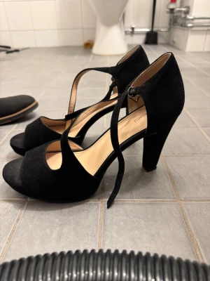 Svarta mocka pumps med klack - Snygga svarta pumps från Dune London i mjuk mocka med rund tå och öppen tå. Skorna har hög klack och smala remmar med spänne som ger en elegant look. Perfekta för dig som gillar stilrena och klassiska detaljer.
