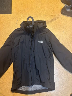 Svart vindjacka från The North Face - Svart vindjacka från The North Face med huva och logga på både bröstet och ryggen. Jackan har dragkedja framtill och meshfoder på insidan. Perfekt för blåsiga dagar. Knappt använd tveka inte på att höra av dig 