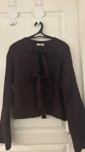 Mörkbrun stickad kofta från Gina Tricot - Mysig vinröd stickad kofta från Gina Tricot med två snygga knytband framtill. Koftan har långärmade, lite bredare ärmar och en ribbad struktur som ger extra vibe. Perfekt för dig som gillar chill och stil på samma gång.