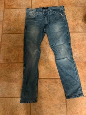Replay Anbass jeans blå, storlek 32 - Snygga Replay Anbass jeans i klassisk blå tvätt med lätt slitningar. Modellen har fem fickor, smal passform och raka ben. Jeansen är tillverkade i mjukt denimtyg med diskreta detaljer på bakfickorna. Perfekt för dig som gillar stilrena och moderna jeans.