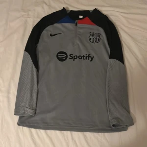 Grå långärmad FC Barcelona tröja - Säljer en grå långärmad FC Barcelona fotbollströja från Nike med svart logga och Spotify-tryck på bröstet. Tröjan har svarta axelpartier, blå och röd detalj vid halsen samt subtilt mönster på ärmarna. Perfekt för dig som vill representera Barca.