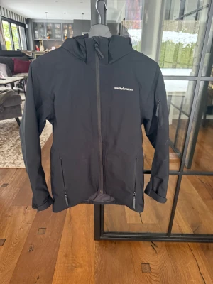 Svart vindjacka från Peak Performance - Svart vindjacka från Peak Performance med huva och dragkedja framtill. Jackan har flera praktiska fickor med dragkedja och en stilren logga på bröstet. Perfekt för blåsiga dagar och enkel att matcha med allt. Snygg och funktionell design.