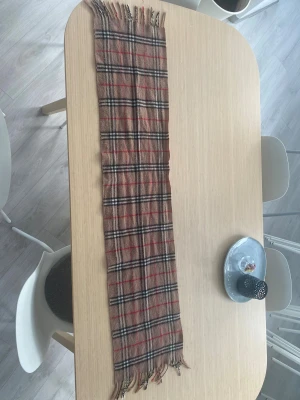 Rutig beige halsduk från Burberry - Snygg klassisk halsduk från Burberry i 100% kashmir. Beige botten med rutigt mönster i svart, vitt och rött. Mjukt material och fransar i ändarna. Perfekt för dig som vill ha en ikonisk accessoar med lyxig känsla och tidlös stil.
