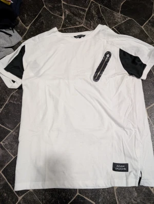 Vit t-shirt med svarta detaljer Adam Vaughn stl L - Snygg vit t-shirt från Adam Vaughn i storlek L med svarta paneler på ärmarna och en cool dragkedjedetalj på bröstet. T-shirten har en modern look och är tillverkad i mjuk bomull. Perfekt för dig som gillar streetstyle och unika detaljer.