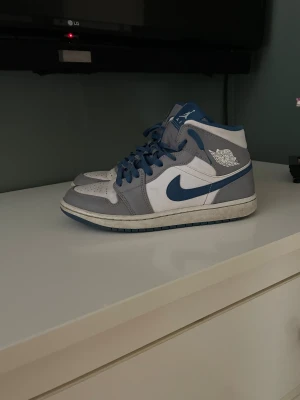 Nike Air Jordan 1 Mid Blå/Grå - Säljer ett par Nike Air Jordan 1 Mid i blått, grått och vitt. Skorna har klassisk Jordan-design med blå swoosh, blå snörning och perforerad tåbox. Ovandelen är i skinn och syntet, med ikonisk Air Jordan-logga på sidan. Perfekta för dig som gillar streetwear och sneakers.