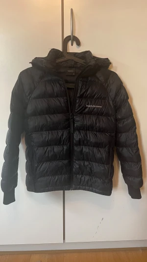 Svart dunjacka från Peak Performance st 150 - Säljer en svart dunjacka från Peak Performance med huva och dragkedja. Jackan är quiltad med breda kanaler, har långa ärmar med muddar och en stilren logga på bröstet. Perfekt för kalla dagar och riktigt snygg till streetwear. Armen har en liten reva som kan enkelt fixas med självjäftande reparstionlsapp. 