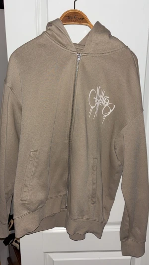 Beige hoodie med Brooklyn-grafik - Säljer en beige hoodie med dragkedja och huva. Framsidan har ett vitt graffiti-tryck och baksidan har en stor Brooklyn-grafik i limegrönt och svart, samt ett foto och text. Hoodien har fickor på sidorna och är långärmad, perfekt för streetwear-stil.