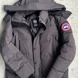 Mörkgrå dunjacka från Canada Goose - Säljer en mörkgrå dunjacka från Canada Goose med ikonisk röd och vit patch på ärmen. Jackan har huva och avtagbar pälsdetalj, samt dragkedja och knappar framtill. Perfekt för kalla vinterdagar och har en clean, stilren look.