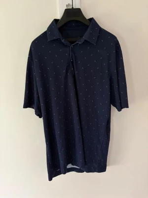 Mörkblå golfpiké från Galvin Green - Snygg mörkblå golfpiké från Galvin Green med diskret mönster och klassisk krage. Skjortan har korta ärmar och små vita detaljer över hela tyget. Perfekt för golfbanan eller en chill dag ute. Storlek: L passar M. Nypris: 999kr