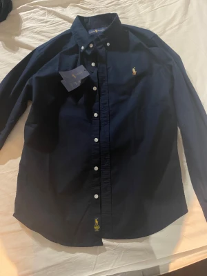 Mörkblå skjorta från Ralph Lauren - Klassisk mörkblå skjorta från Ralph Lauren med broderad gul logga på bröstet och knappar framtill. Skjortan har lång ärm, button-down krage och är tillverkad i bomull. Perfekt för en clean och stilren look.