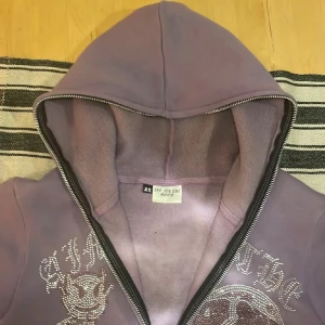 Ljuslila hoodie med strassmotiv XS - Säljer en ljuslila hoodie från See You In The Moon i storlek XS. Tröjan har huva, dragkedja och är fodrad med mjuk fleece. På framsidan finns ett stort strassmotiv med text och figur, vilket ger en unik och glittrig vibe. Perfekt för dig som gillar statement-plagg.