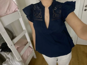 Mörkblå blus med volangärm och broderi - Säljer en mörkblå blus med volangärmar och snygga broderade detaljer upptill. Blusen har en djup v-ringning och är ärmlös med en luftig passform. Perfekt för dig som gillar feminina och trendiga plagg.