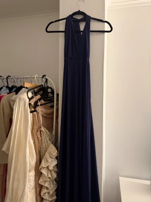 Marinblå långklänning  - Snygg marinblå långklänning med knute upptill och djup v-ringning. Klänningen har breda axelband och öppen rygg med resår i midjan för en bekväm passform. Tillverkad i ett mjukt, följsamt material som faller fint.
