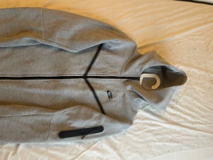 Grå zip-hoodie från Nike med huva - Snygg grå hoodie från Nike med hel dragkedja och svart logga på bröstet. tjocktröjan har huva, svarta detaljer längs dragkedjan och en ficka med dragkedja på ärmen. Perfekt för en sportig och avslappnad stil.