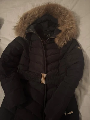 Svart pufferjacka med päls från Hollies - Snygg svart pufferjacka från Hollies med fluffig beige fuskpäls på huvan och guldfärgat bälte i midjan. Jackan har dragkedja framtill, långa ärmar och quiltad design. Perfekt för kalla dagar när du vill vara både varm och stilren. Äkta