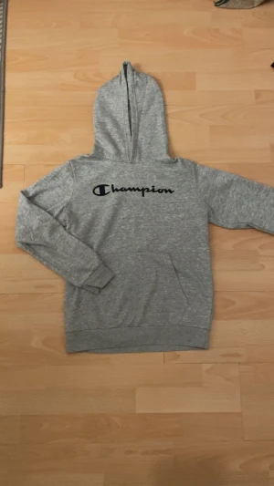 Grå hoodie från Champion XL - Grå hoodie från Champion i storlek XL med svart logotyp tryckt över bröstet. Tröjan har huva, magficka och långa ärmar. Mjuk insida och klassisk passform, perfekt för chill dagar eller när du vill ha en enkel streetstyle-look. Använd ett få par gånger!