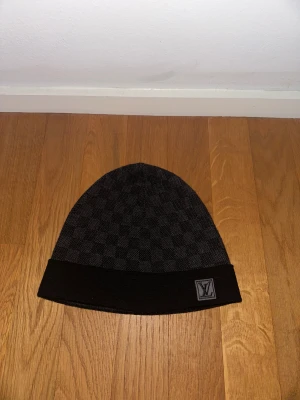 Svart Louis Vuitton mössa - Exklusiv svart mössa från Louis Vuitton med klassiskt Damier-rutmönster och LV-logga framtill. Mössan har en uppvikt kant och är tillverkad i mjukt material som passar perfekt för kyliga dagar. Levereras med originalbox och certifikat.