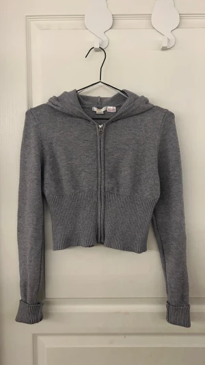 Grå croppad hoodie från Gina Tricot - Grå croppad hoodie från Gina Tricot med ribbad nederkant och muddar. Tröjan har huva, dragkedja framtill och är långärmad. Perfekt för dig som gillar en chill och trendig look. Materialet är mjukt och stickat för extra komfort.