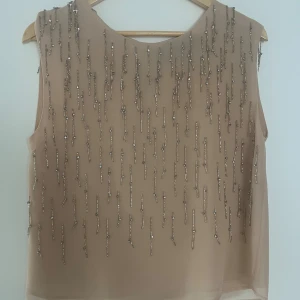 Beige blus med pärlor från Patrizia Pepe - Säljer en beige blus från Patrizia Pepe i polyester med coola vertikala pärldetaljer och paljetter framtill. Blusen är ärmlös och har en rund hals samt knapp i nacken. Perfekt för dig som gillar glittriga detaljer och vill sticka ut lite extra.