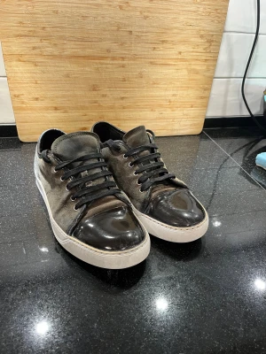 Gråsvarta sneakers med mocka och lack - Snygga sneakers i mocka med svarta lackdetaljer på tå och häl. Skorna har svarta snören, vit sula och en stilren, rund tå. Perfekta för dig som gillar en mix av material och en urban look. Sulan och sidorna är målade, och mockan är sliten, annars är de bra skick.