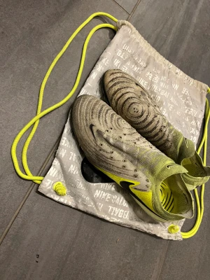 Nike Phantom GT2 fotbollsskor gul/svart - Säljer ett par Nike Phantom GT2 fotbollsskor i grått och neongult med svarta detaljer. Skorna har ett mönster med cirklar på ovandelen, strumpliknande skaft och plös, samt dobbar för bra grepp på planen. Snygg och modern design, perfekt för dig som vill sticka ut på fotbollsplanen. Skorna har några dobbar som har slitits ut (bilderna ovanför) annars bra skick. Låda följs med givitvis
