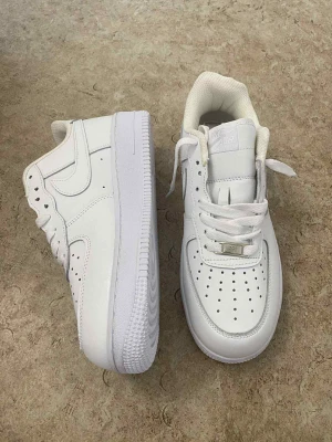 Nike Air Force 1 vita sneakers - Klassiska Nike Air Force 1 sneakers i helvitt skinn med perforerad tå, rund tå och platt sula. Snygg och tidlös modell med snörning och diskret Nike-logga på sidan. Perfekta för dig som vill ha en clean och stilren look.