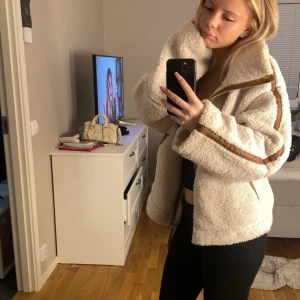 Beige teddyjacka med bruna detaljer - Säljer en supermjuk och oversized teddyjacka i beige med bruna detaljer längs dragkedjan och ärmarna. Jackan har hög krage, stora fickor och grov dragkedja. Perfekt för kalla dagar och ger en chill vibe till outfiten. Köpt för 900