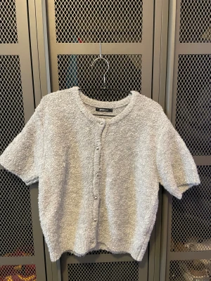 Grå kortärmad kofta från Gina Tricot - Mysig grå kofta från Gina Tricot med korta ärmar och rund hals. Koftan har en fluffig, stickad struktur och knäpps med silvriga knappar framtill. Perfekt för dig som gillar en chill och trendig look.