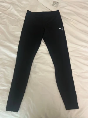 Svarta träningsleggings från Puma - Svarta långleggings från Puma med vit logga på höften. De är tillverkade i ett syntetmaterial med dryCELL-teknologi som håller dig torr under träningen. Tight passform och dragkedja vid benslut för extra smidighet. Perfekta för löpning eller gym.