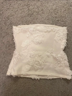 Vit spetsprydd tubtopp från BikBok - Supersöt vit tubtopp från BikBok med 3D-blommiga spetsdetaljer och dragkedja i storlek S. Har gummiband i övre kant så tröjan sitter på plats. TOPPEN ÄR OAVNÄND, Orginalpris 299kr
