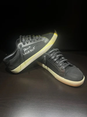 Svarta Saint Laurent sneakers - Svarta sneakers från Saint Laurent med vit text på sidan och Paris-logga på innersulan. Skorna har rund tå, snörning och en beige platt sula med fiskbensmönster undertill. Ovandelen är i canvas och detaljer i skinn, vilket ger en clean och stilren vibe.