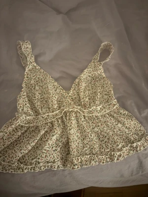 Blommig volang linnetopp med v-ringning - Supersöt linnetopp med blommigt mönster i ljusa toner, v-ringad framtill och breda axelband. Toppen har volangdetaljer och smock baktill för skön passform. Perfekt för varma dagar och ger en romantisk vibe till din outfit.