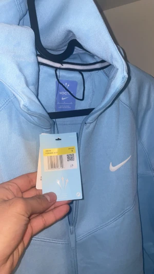 Nike NOCTA ljusblå hoodiejacka S - Nike NOCTA hoodiejacka i ljusblått med dragkedja, huva och vita loggor på bröstet och ärmen. Jackan har en clean look med snygga sömdetaljer och är tillverkad i mjuk bomullsmix. Perfekt för dig som gillar streetwear och vill sticka ut med en fräsch färg.