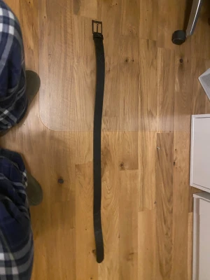 Svart skärp från Guess med logomönster - Snyggt svart bälte från Guess med präglat logomönster längs hela remmen och metallspänne. Bältet har en klassisk rektangulär form och Guess-triangel vid spännet. Perfekt accessoar för att lyfta din outfit.